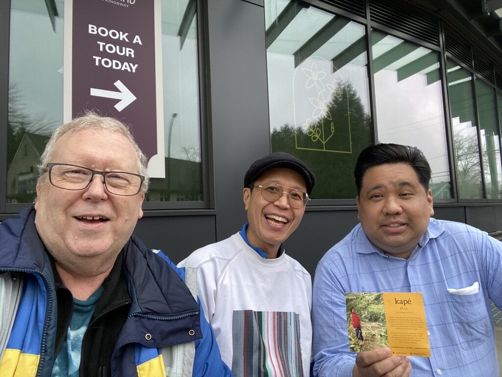 Joe Planta, Dale McGladdery and Roland Tanglao on Kingsway in Vancouver just east of Analog Coffee at an outdoor pseudo-patio. Covered! Joe is holding up a marketing postcard for kapé which is a social venture promoting ethically sourced, farmer-first, direct trade coffee from Philippine Indigenous growers and batch roasted on unceded Coast Salish Territories (Vancouver, Canada).